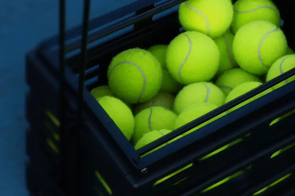 Une caisse noire remplie de balles de tennis jaune vif sur une surface bleue. La texture des balles duveteuses contraste avec la caisse lisse, créant une ambiance sportive