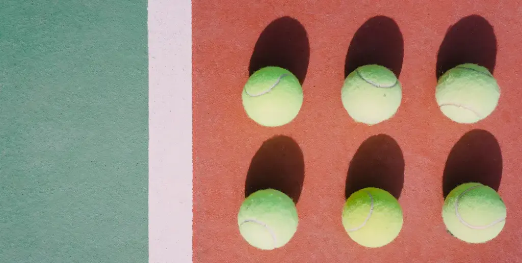 Six balles de tennis disposées en deux rangées sur un court rouge. Les ombres sont projetées vers la droite, avec un bord rayé vert et blanc à gauche, créant un contraste